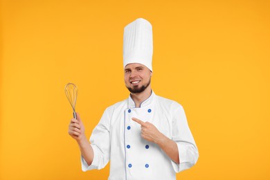 Photo of Happy professional confectioner in uniform pointing at whisk on yellow background