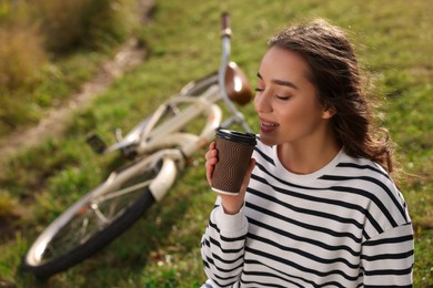Young woman sitting on green grass and drinking coffee near bicycle outdoors, space for text