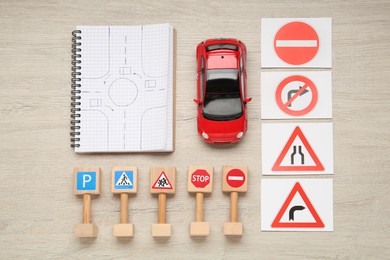 Photo of Many different road signs, notebook with sketch of roundabout and toy car on wooden table, flat lay. Driving school
