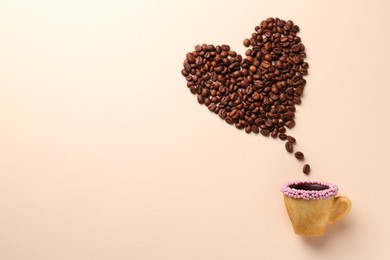 Photo of Coffee beans spilled from edible biscuit cup in shape of heart on beige background, top view. Space for text