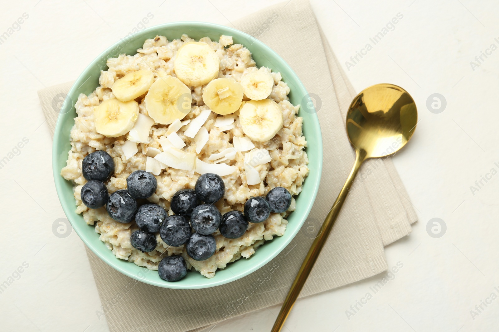 Photo of Tasty oatmeal with banana, blueberries, coconut flakes and honey served in bowl on beige table, top view