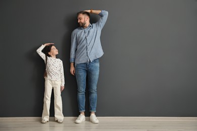 Photo of Father and daughter comparing their heights near black wall indoors, space for text