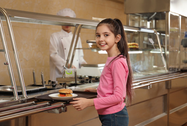 Photo of Little girl with plastic tray and burger near serving line in canteen. School food