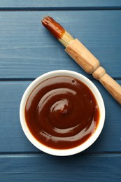 Photo of Tasty barbeque sauce in bowl and brush on blue wooden table, top view