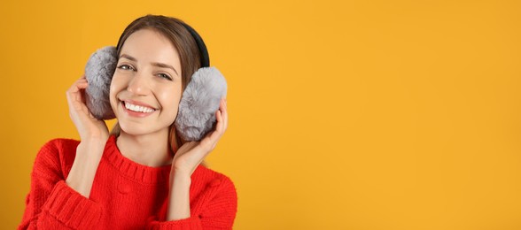 Happy woman wearing warm earmuffs on yellow background, space for text