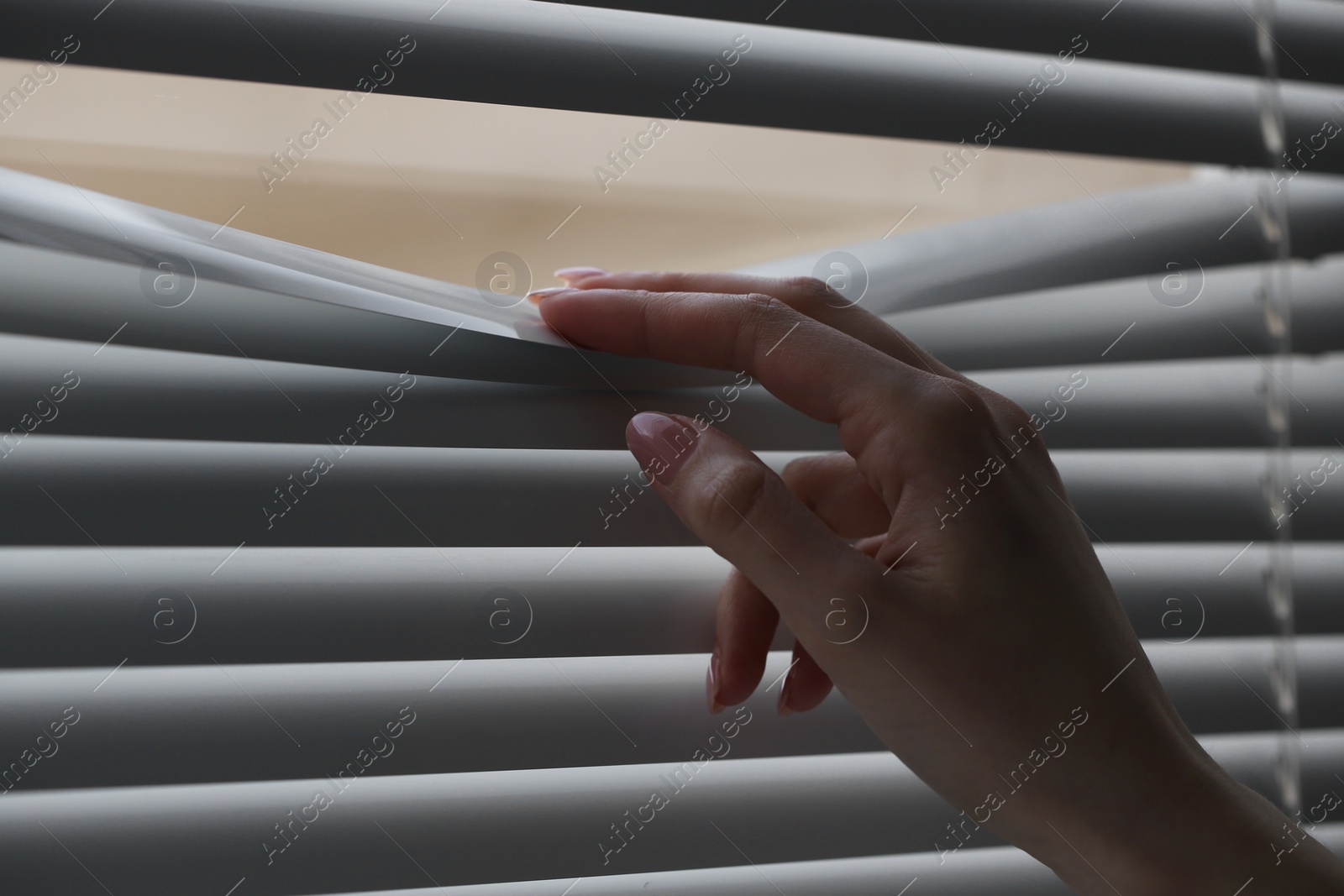 Photo of Woman separating slats of white blinds indoors, closeup