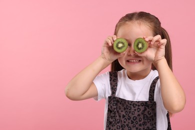 Photo of Smiling girl covering eyes with halves of fresh kiwi on pink background, space for text