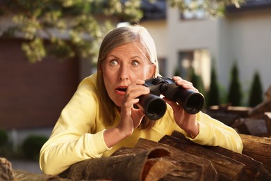Photo of Concept of private life. Curious senior woman with binoculars spying on neighbours over firewood outdoors