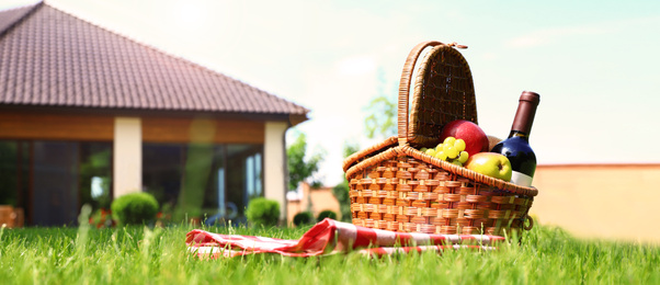 Image of Picnic basket with fruits, bottle of wine and checkered blanket on green grass in garden, space for text. Banner design