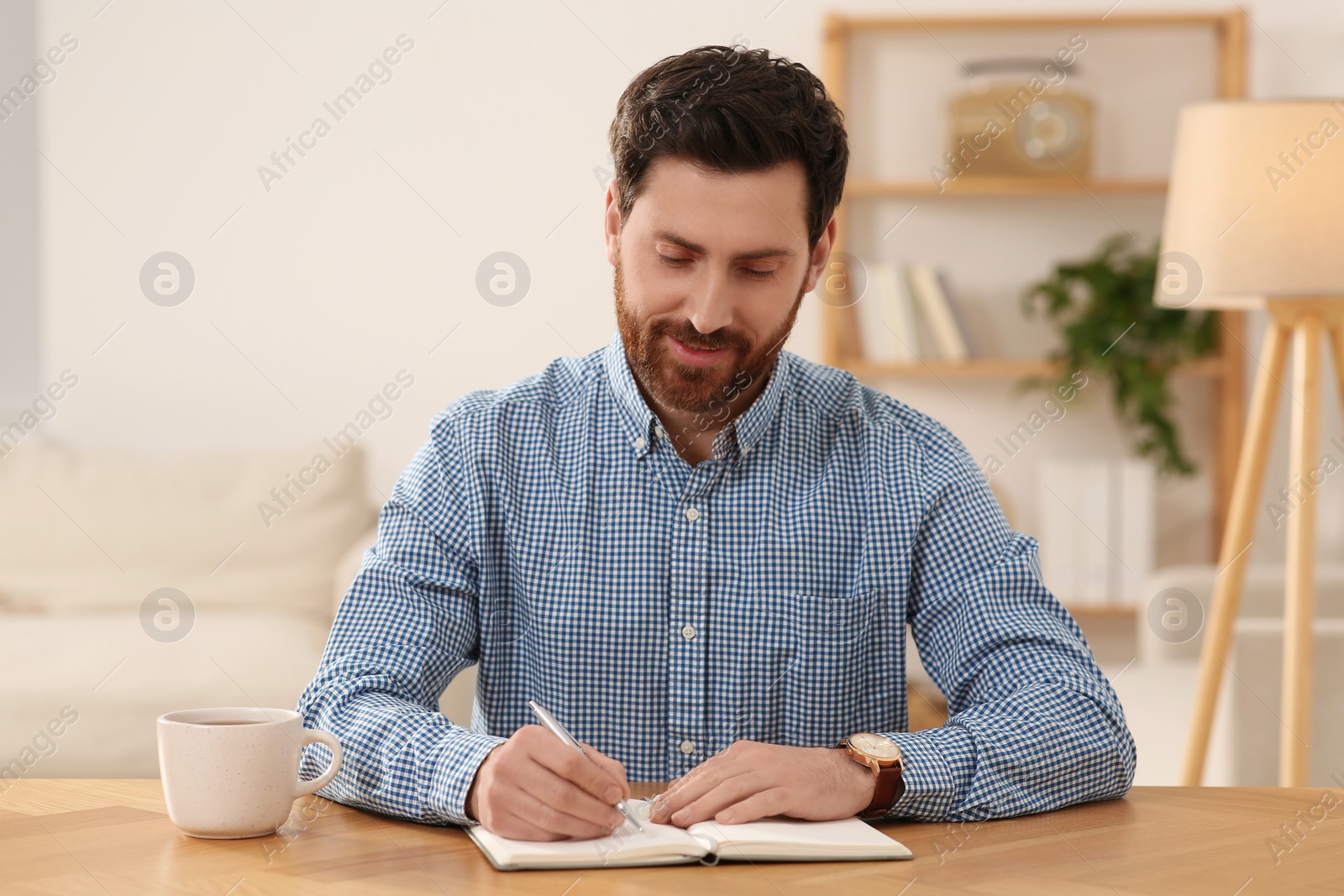 Photo of Happy man taking notes during video call at wooden desk indoors, view from camera