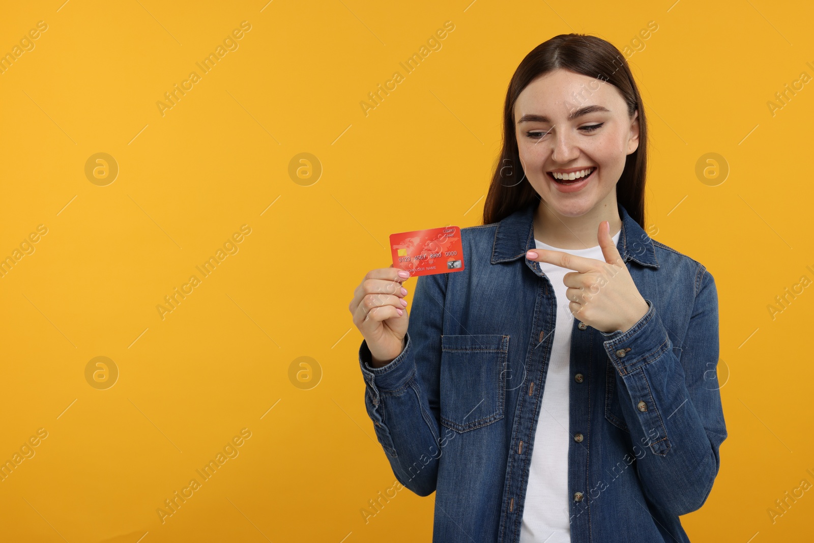 Photo of Happy woman pointing at credit card on orange background, space for text