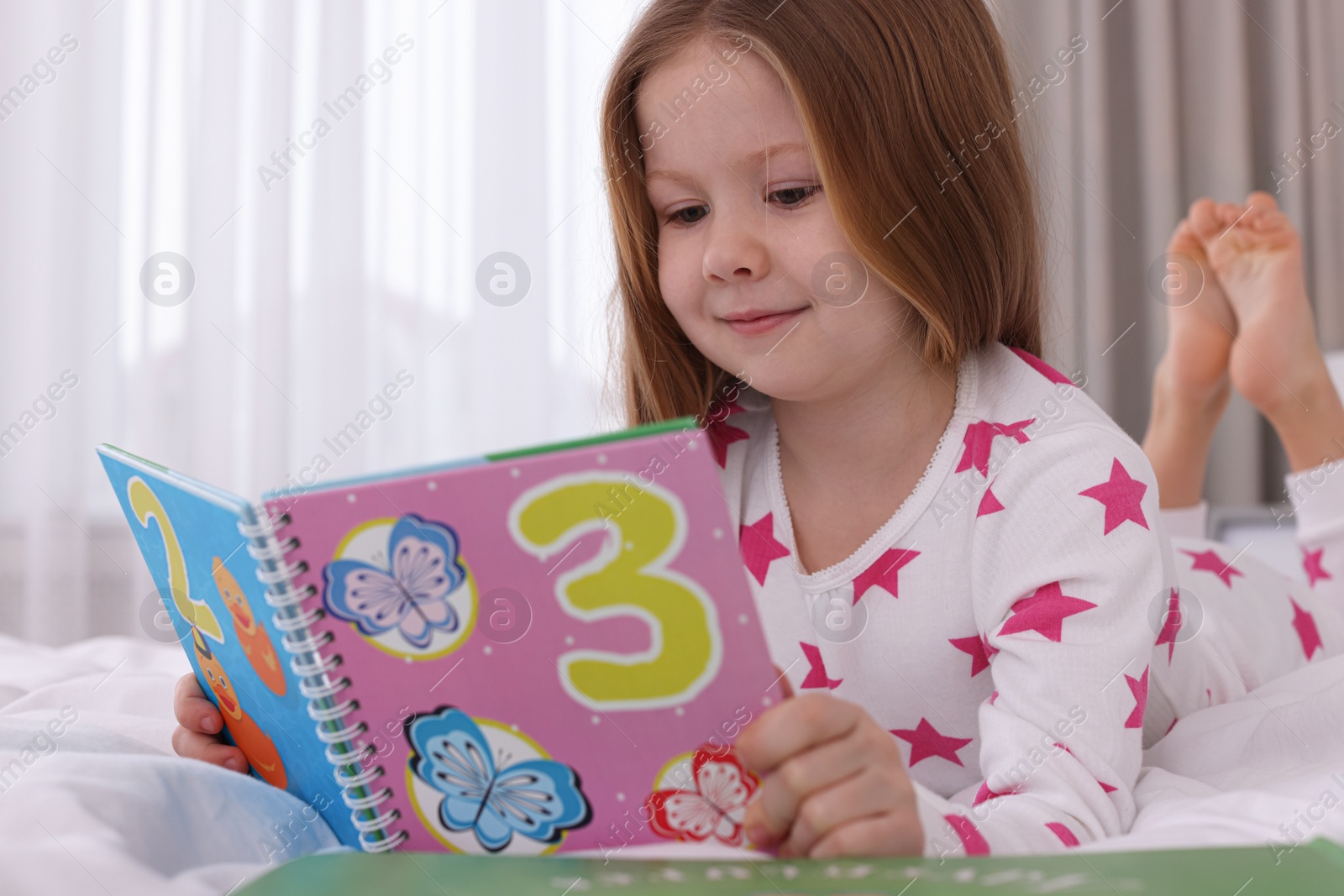 Photo of Cute little girl reading book on bed at home