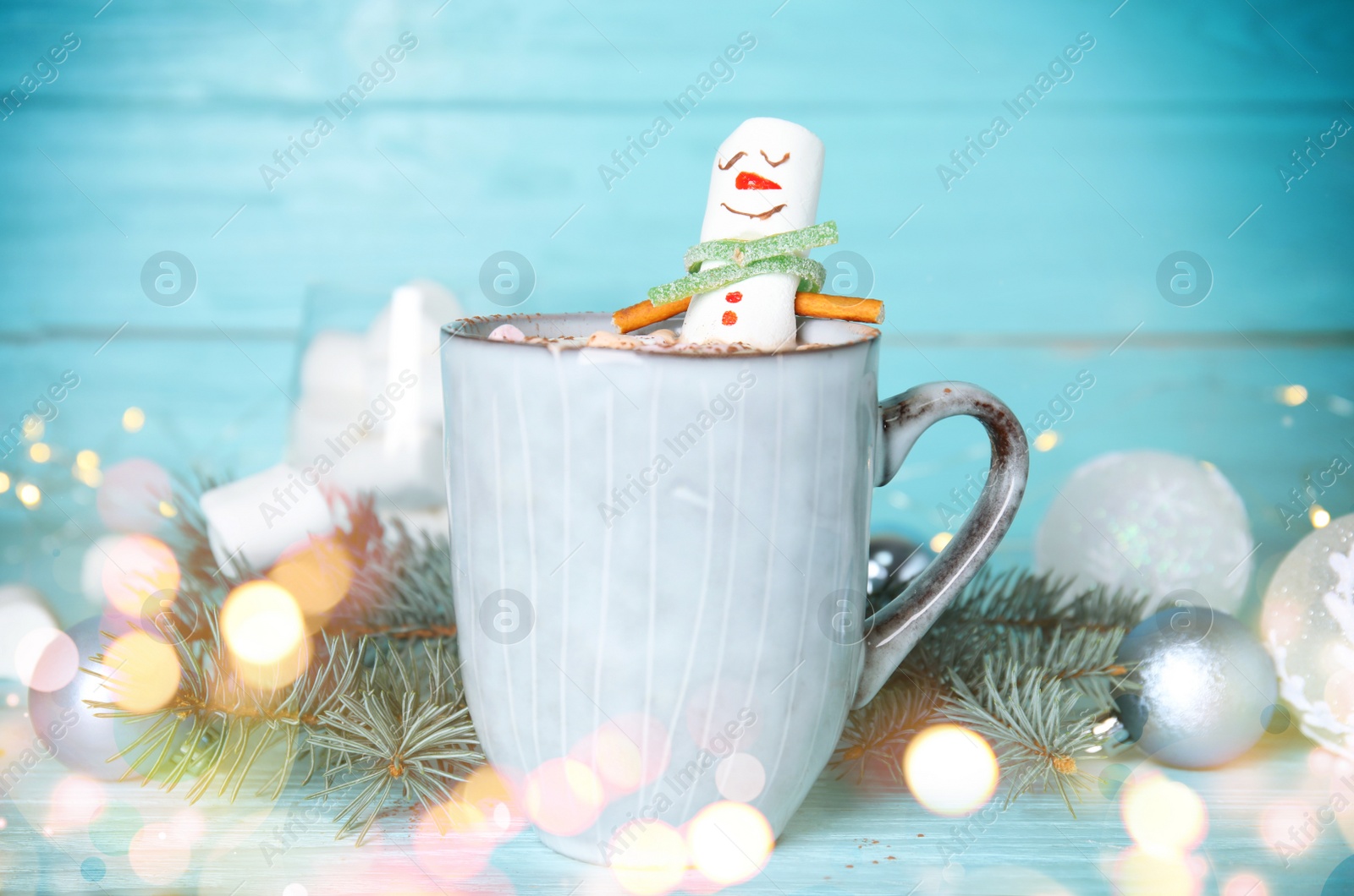 Image of Funny marshmallow snowman in cup of hot drink on light blue wooden table. Bokeh effect
