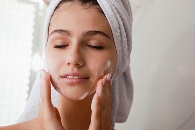 Photo of Beautiful teenage girl applying cleansing foam onto face in bathroom, closeup. Skin care cosmetic