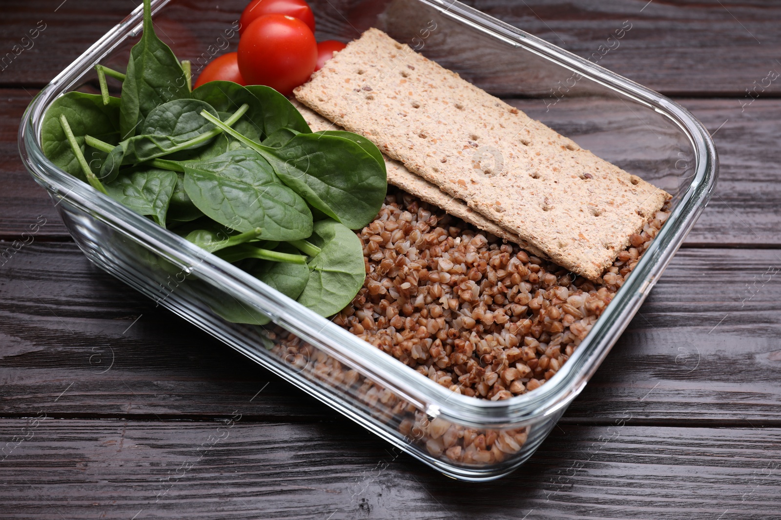 Photo of Glass container with buckwheat, fresh tomato, spinach and crispbreads on wooden table, closeup