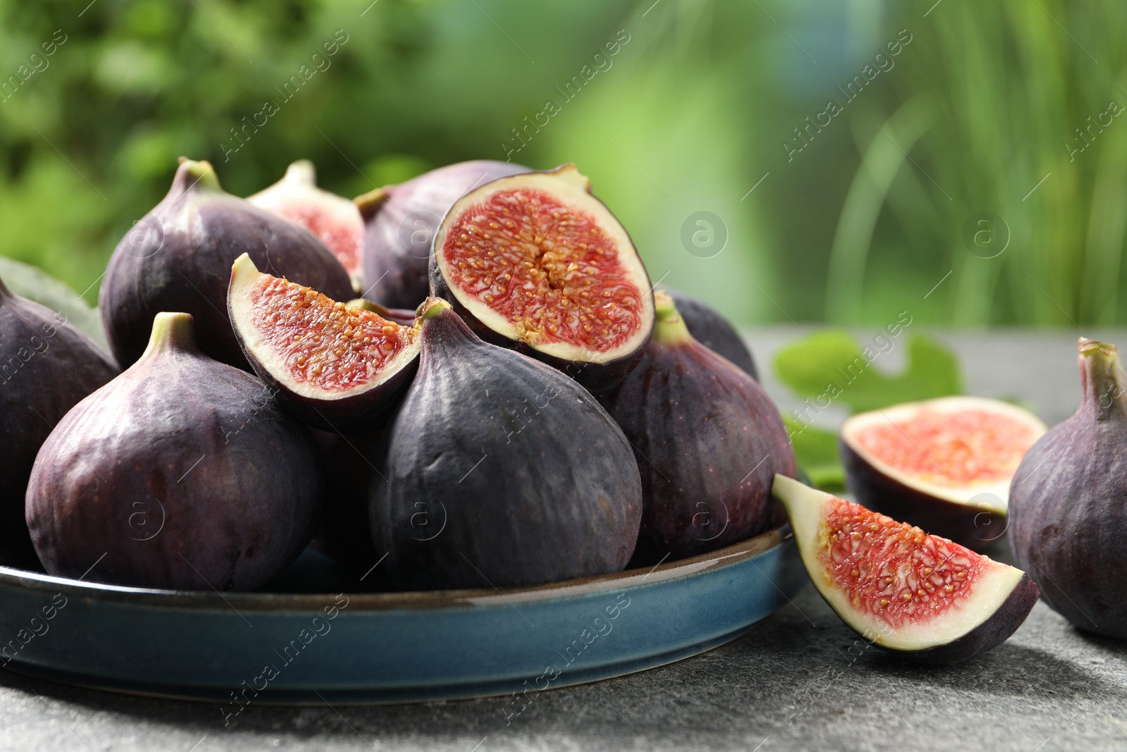 Photo of Whole and cut ripe figs on light grey textured table against blurred green background, closeup