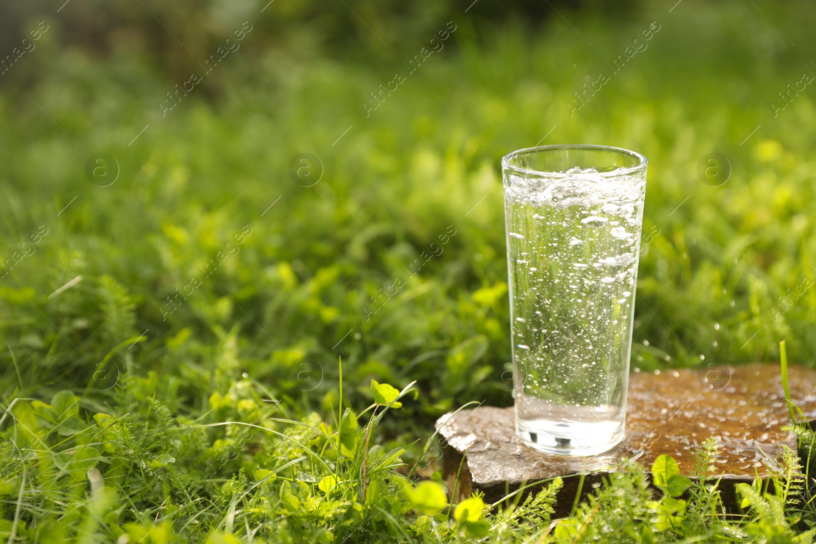 Photo of Glass of fresh water on stone in green grass outdoors. Space for text