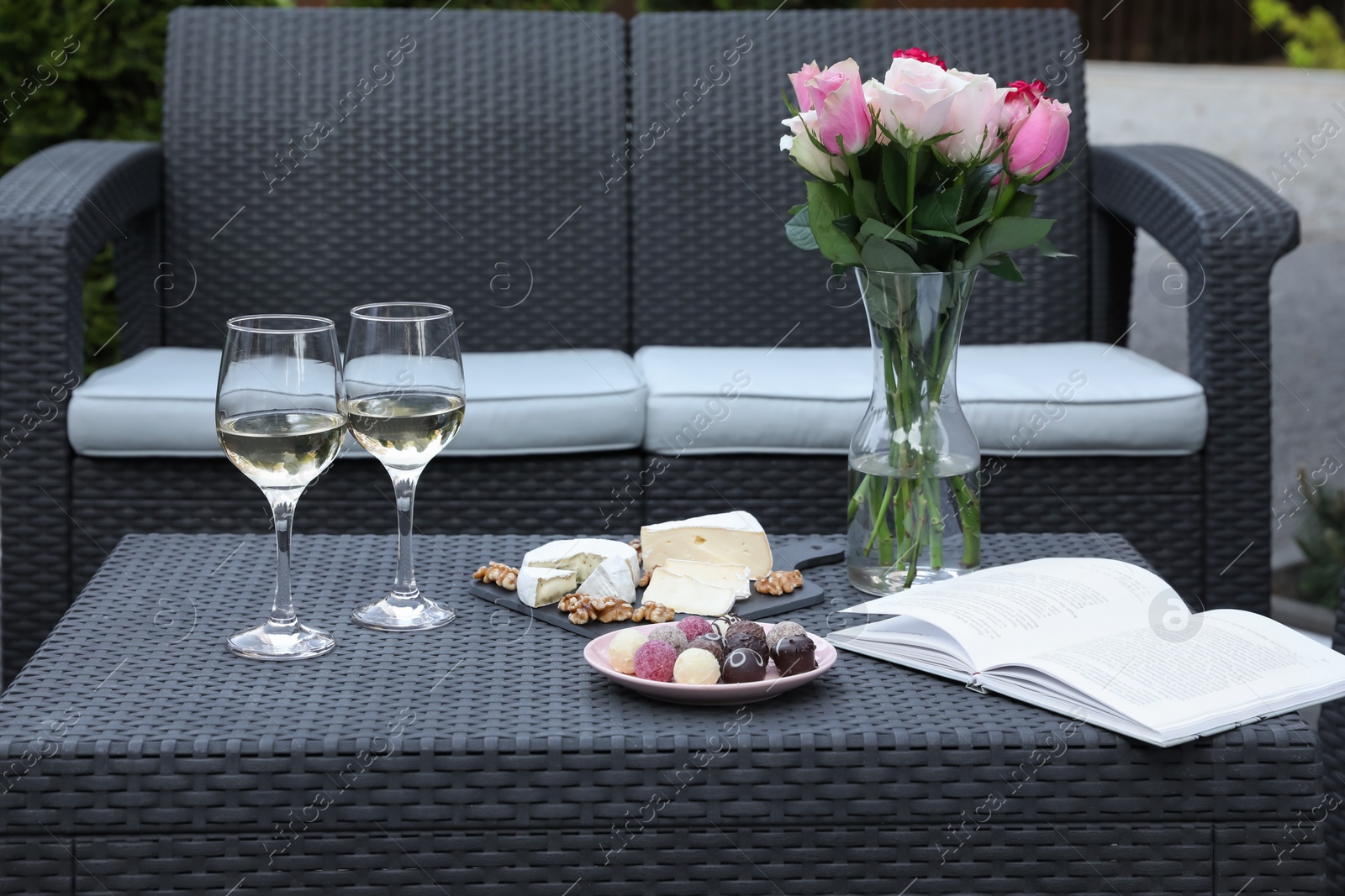 Photo of Vase with roses, glasses of wine, open book and different snacks on rattan table on outdoor terrace