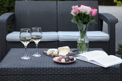 Photo of Vase with roses, glasses of wine, open book and different snacks on rattan table on outdoor terrace