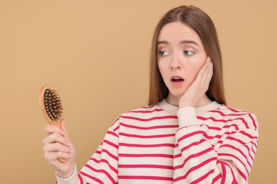 Photo of Emotional woman holding brush with lost hair on beige background. Alopecia problem
