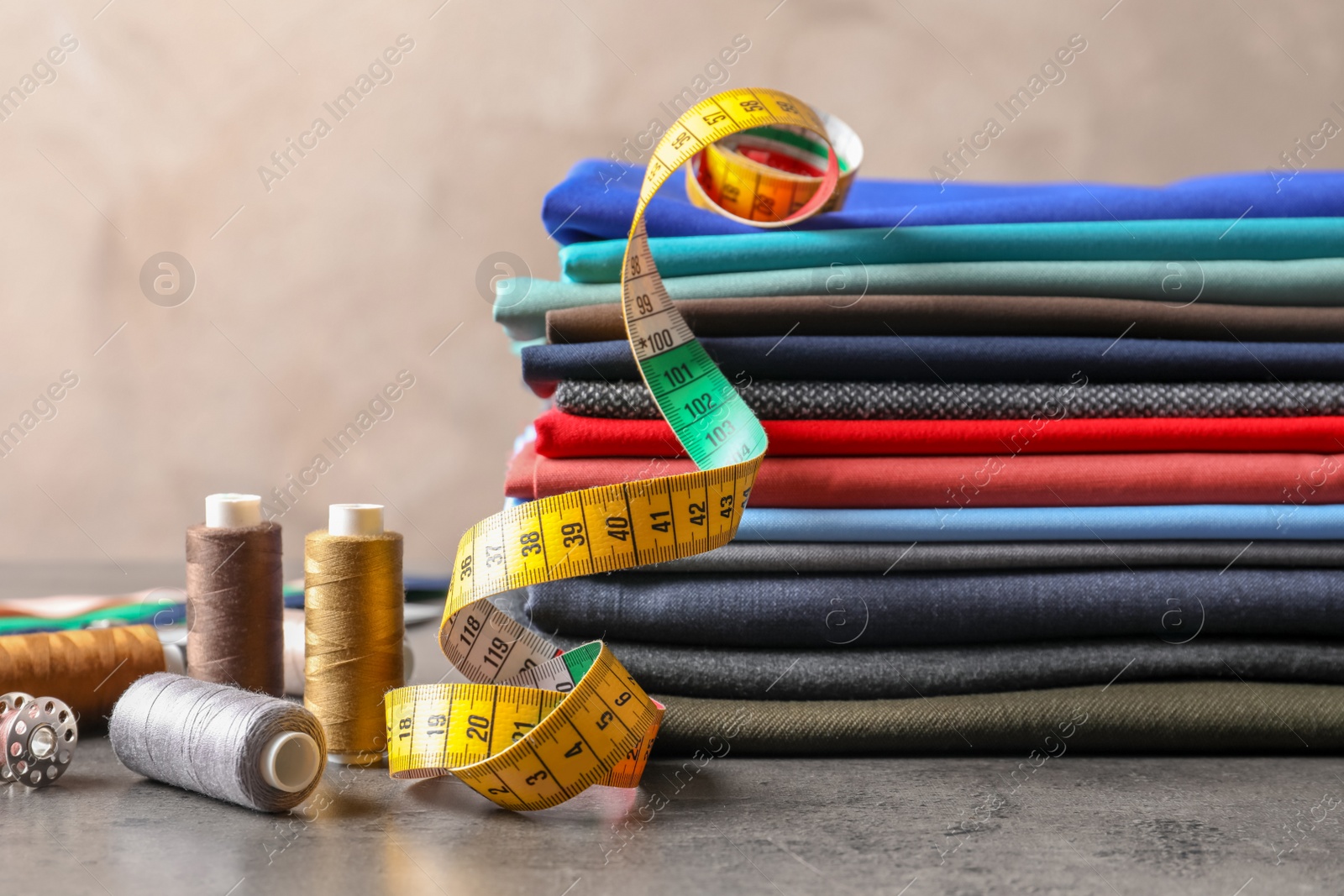 Photo of Bobbins with threads, measuring tape and stack of colorful fabrics on table. Tailoring accessories