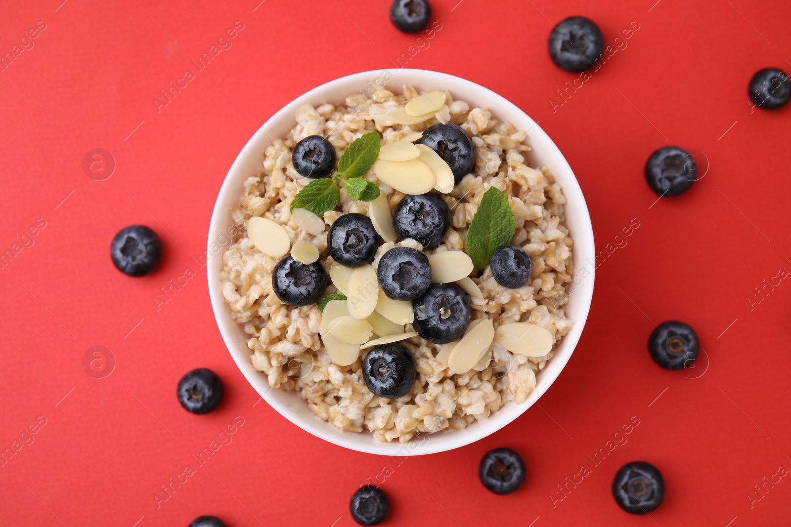 Photo of Tasty oatmeal with blueberries, mint and almond petals in bowl surrounded by fresh berries on red background, flat lay