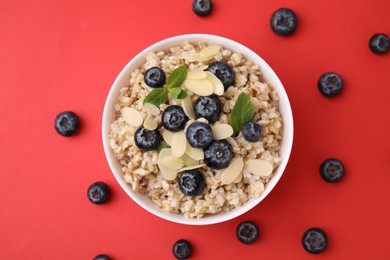 Photo of Tasty oatmeal with blueberries, mint and almond petals in bowl surrounded by fresh berries on red background, flat lay