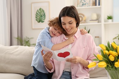 Little son congratulating his mom with Mother`s day at home. Woman holding handmade greeting card