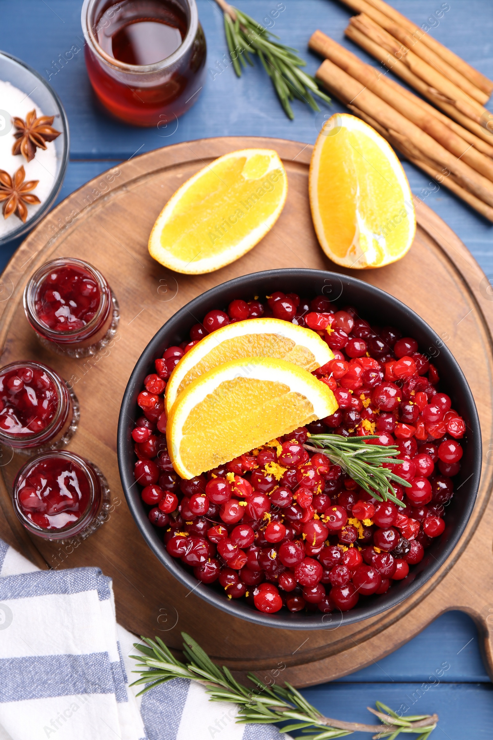 Photo of Cranberries in bowl, jars with sauce and ingredients on blue wooden table, flat lay