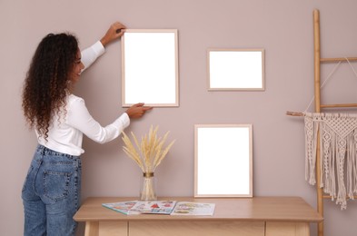 Photo of African American woman hanging empty frame on pale rose wall over table in room. Mockup for design