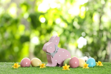 Cute wooden Easter bunny and dyed eggs on green grass against blurred background