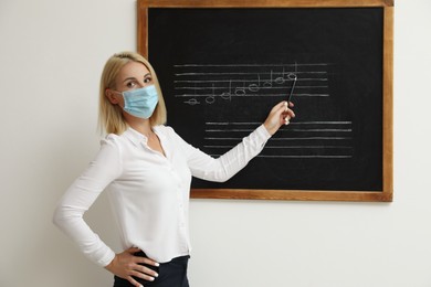 Photo of Teacher wearing protective mask near black chalkboard with music notes in classroom