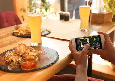 Photo of Woman playing game using smartphone at table with tasty BBQ wings in cafe, closeup
