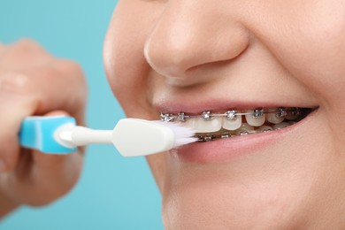 Smiling woman with dental braces cleaning teeth on light blue background, closeup