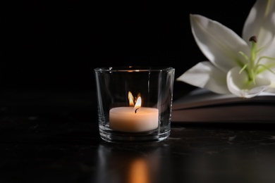 Burning candle, book and white lily on table in darkness. Funeral symbol