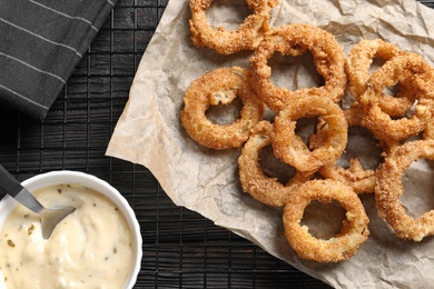 Photo of Cooling rack with homemade crunchy fried onion rings and sauce on wooden background, top view