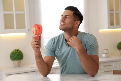 Photo of Man enjoying air flow from portable fan at table in kitchen. Summer heat