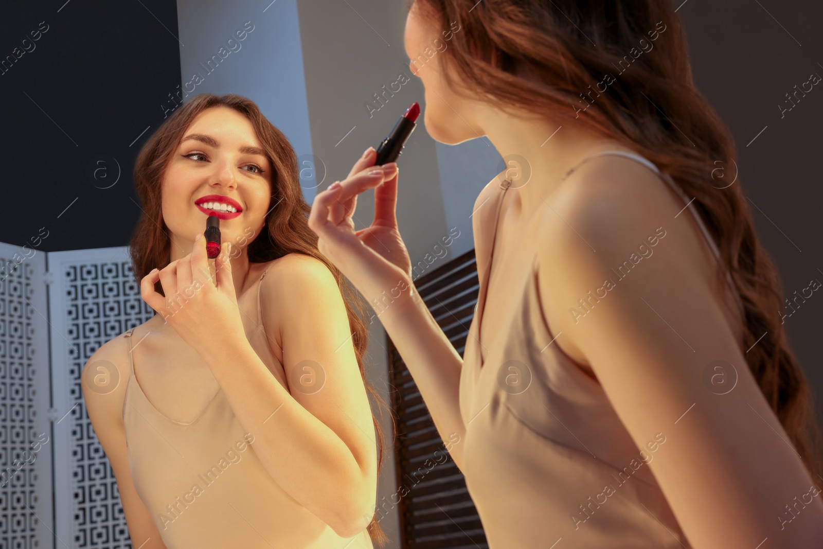 Photo of Beautiful young woman in elegant dress applying lipstick near mirror indoors