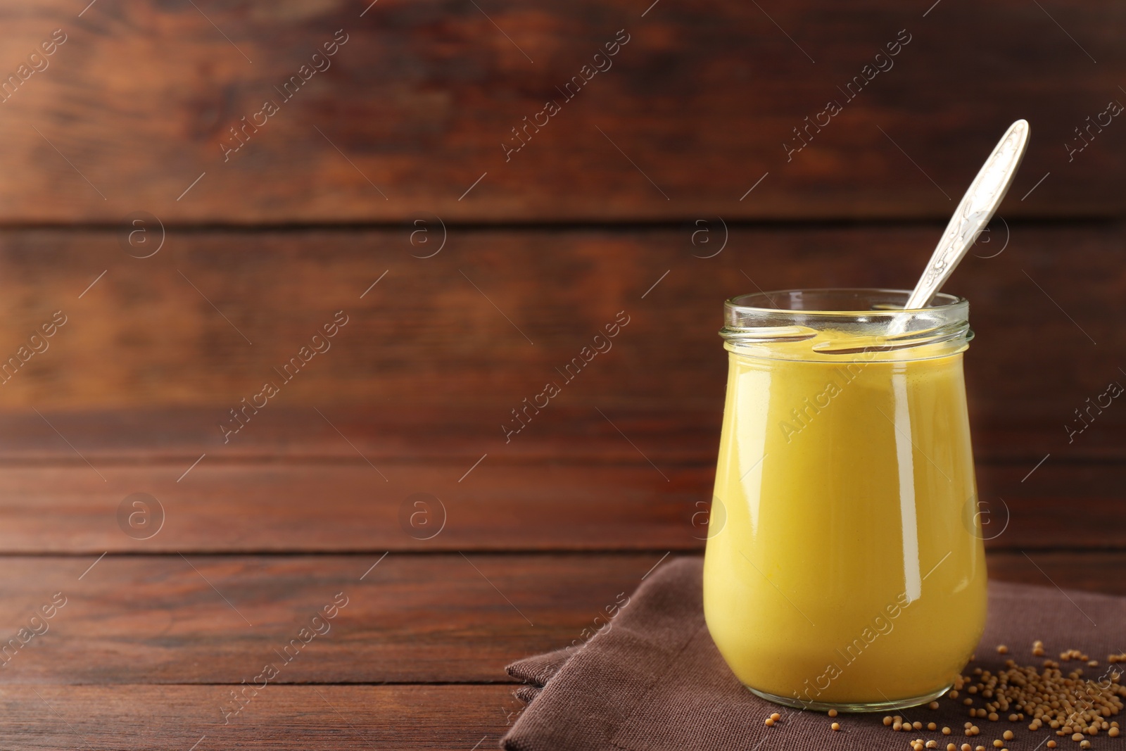 Photo of Tasty mustard sauce and spoon in glass jar with dry seeds on wooden table, space for text