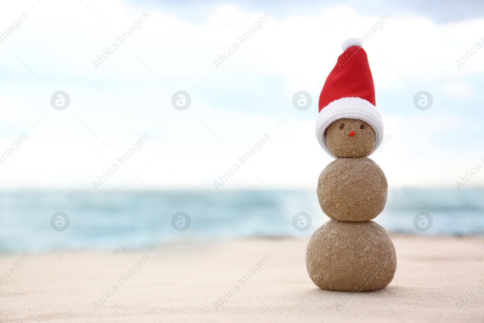 Photo of Snowman made of sand with Santa hat on beach near sea, space for text. Christmas vacation