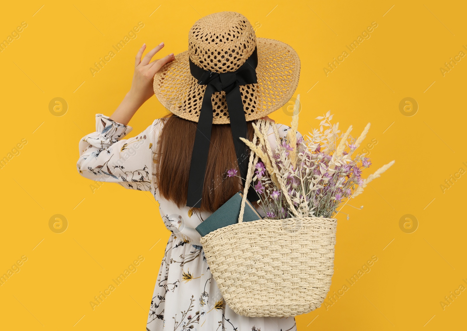 Photo of Woman holding beach bag with beautiful bouquet of wildflowers and book on yellow background, back view