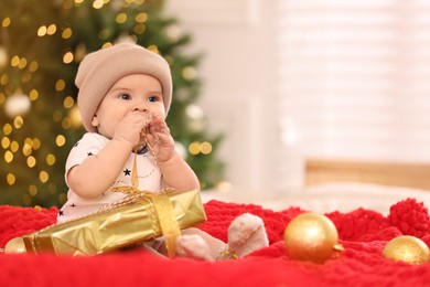 Photo of Cute little baby with Christmas gift on red blanket against blurred festive lights, space for text. Winter holiday