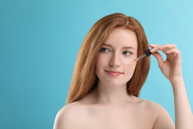 Beautiful woman with freckles applying cosmetic serum onto her face against light blue background. Space for text