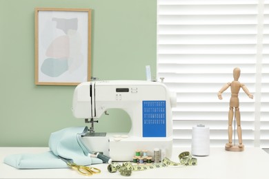 Photo of Modern sewing machine with cloth and craft accessories on white table near window in room