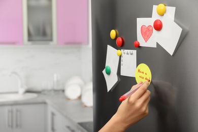 Photo of Woman writing message on note stuck to refrigerator door at home, closeup