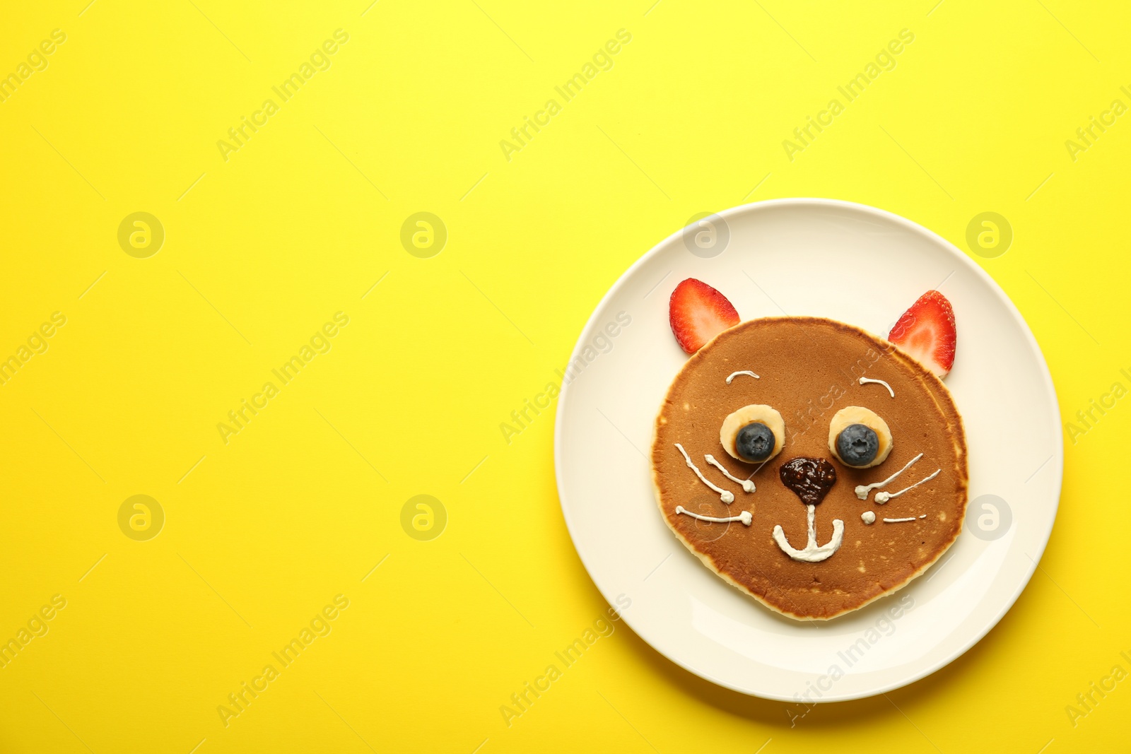 Photo of Creative serving for kids. Plate with cute cat made of pancakes, berries, cream, banana and chocolate paste on yellow background, top view. Space for text