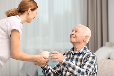 Photo of Elderly man taking cup of tea from female caregiver at home