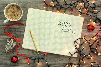 Photo of Inscription 2022 Goals written in planner, cup of coffee and Christmas decor on wooden background, flat lay. New Year aims
