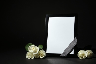 Funeral photo frame with ribbon and white roses on dark table against black background. Space for design
