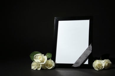 Photo of Funeral photo frame with ribbon and white roses on dark table against black background. Space for design